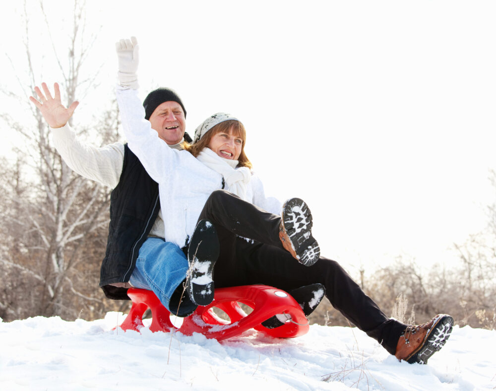 Older married couple playing in the snow, participating in one of our winter date ideas; they are sledding down a hill