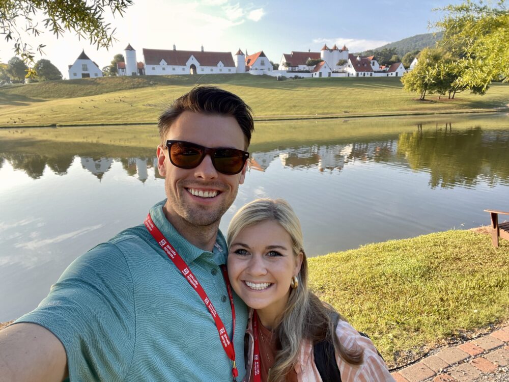 a happy married couple taking a selfie in front of a lake and beautiful buildings at a winshape marriage retreat