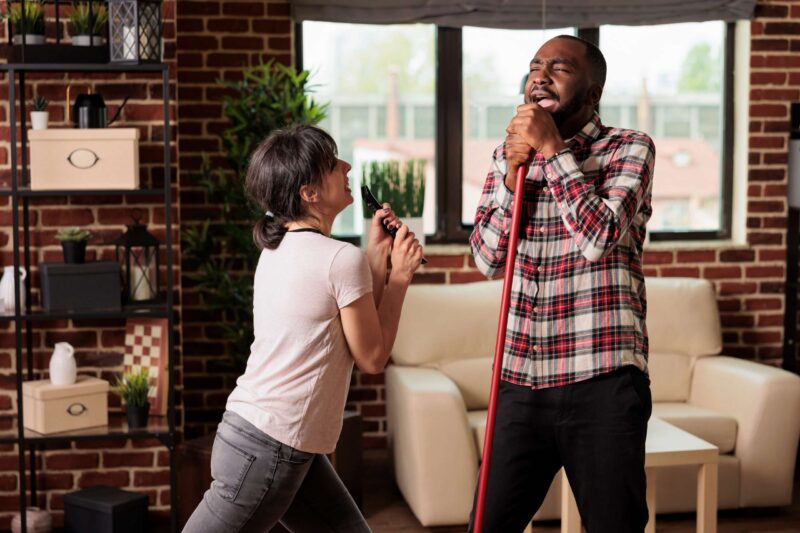 A young married couple enjoying one of our spring date ideas, spring cleaning, they're singing together using the TV remote and broom as microphones