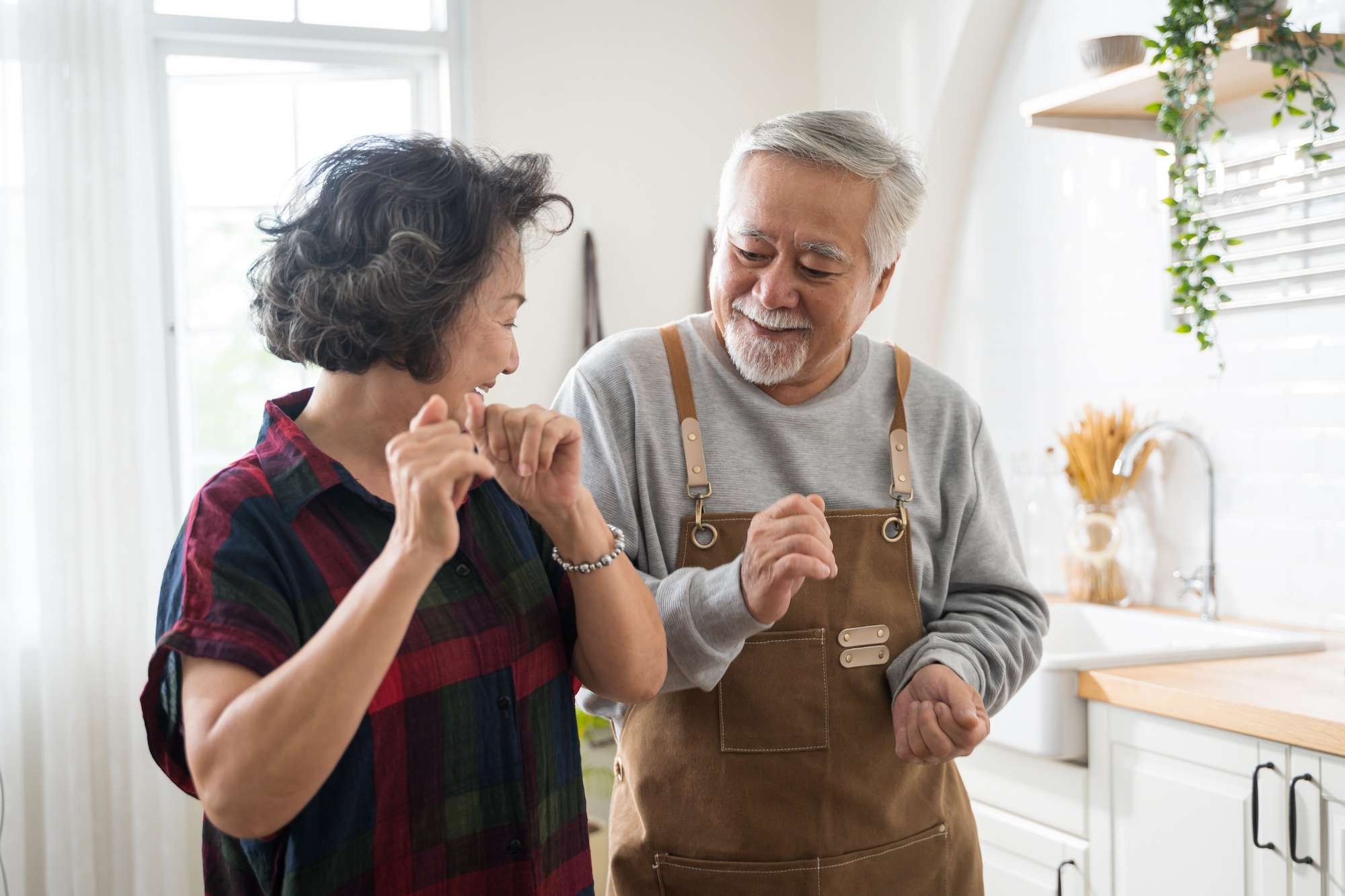 Asian mature senior couple dancing and smiling in kitchen at home.