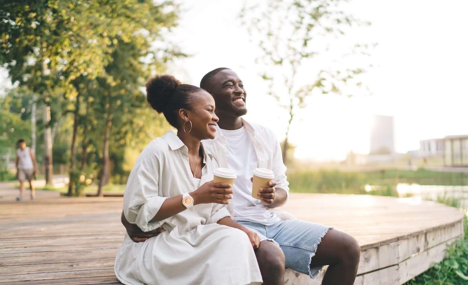 Smiling African American man and woman sitting together with coffee cups while enjoying weekend near lake in park against green trees